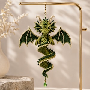May include: A crocheted green dragon ornament with textured scales, large wings, and two horns. The dragon has a long, spiraling tail that ends with a small green teardrop-shaped bead. It is suspended from a gold-colored metal display stand.