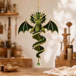 May include: A crocheted green dragon ornament with a spiraling tail, textured scales, and large wings. The dragon features cream-colored horns and a small green crystal bead hanging from the tip of its tail, suspended by a gold-toned metal chain.