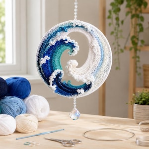 May include: A circular crochet wall hanging featuring a stylized ocean wave design in shades of deep blue, teal, and white. The piece is suspended by a clear beaded string and features a faceted crystal drop at the bottom center.
