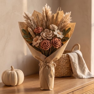 May include: A bouquet of crocheted flowers in shades of rust, cream, and beige, featuring roses and daisies mixed with dried pampas grass and wheat stalks. The arrangement is wrapped in brown paper and tied with a simple twine bow at the base.
