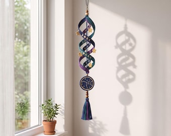 May include: A hanging spiral mobile featuring a gradient of teal, blue, and purple fabric with gold moon phase and star accents. A circular pendant with a gold constellation design hangs below, finished with a multi-colored tassel and wooden beads.