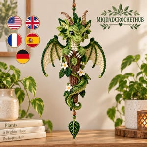 Può includere: Una decorazione da appendere all'uncinetto a forma di drago verde con grandi occhi, corna e ali, avvolto attorno a un ramo marrone testurizzato decorato con fiori bianchi e foglie verdi. Il testo MIQDADCROCHETHUB è visibile in alto a destra.