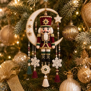 May include: A crocheted nutcracker ornament centered on a crescent moon, featuring a maroon uniform with gold trim, a tall hat, and a white beard. The piece is adorned with hanging white snowflake motifs, small pearl-like beads, and colorful tassels.