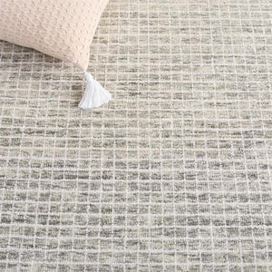 May include: A textured area rug featuring a grid pattern in shades of cream, beige, and charcoal gray. A corner of a light beige woven pillow with a white tassel is visible in the upper left, resting on the surface of the patterned rug.