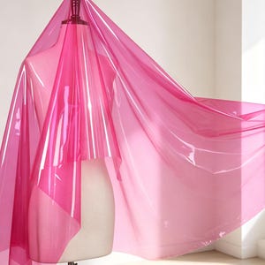 May include: A translucent, vibrant hot pink vinyl sheet draped over a neutral-colored dress form. The material is glossy, smooth, and flexible, creating soft folds and light reflections as it extends outward from the mannequin's shoulder.