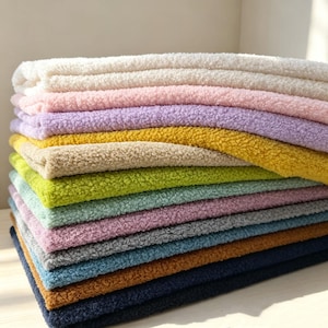 Könnte beinhalten: Ein Stapel gefalteter Stoffstücke mit einer texturierten, lockigen, sherpa-artigen Oberfläche. Die Schichten weisen eine Vielzahl von Farben auf, darunter Weiß, Hellrosa, Lavendel, Gelb, Beige, Limettengrün, Mint, Mauve, Grau, Blau, Braun und Marineblau.
