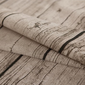Può includere: Un pezzo di tessuto con un motivo stampato a venature del legno in tonalità marrone chiaro, beige e marrone scuro. Il materiale è ripiegato su se stesso, mostrando una texture realistica di assi di legno con nodi e venature verticali.