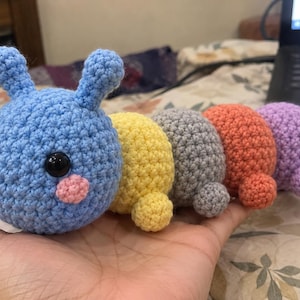 May include: A multi-colored crochet caterpillar toy featuring a large blue head with two antennae, black safety eyes, and pink cheeks. The body consists of four rounded segments in yellow, gray, orange, and purple, each with a small matching foot.
