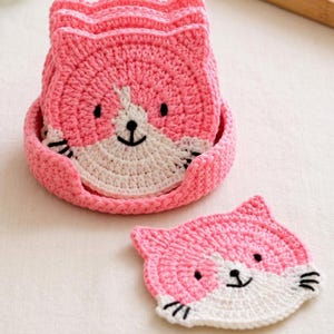 Könnte beinhalten: Ein Set aus gehäkelten, katzenförmigen Untersetzern in Rosa und Weiß, die in einem passenden gehäkelten Halter in Rosa gestapelt sind. Jeder Untersetzer zeigt ein rosa Gesicht mit weißen Details, schwarzen gestickten Augen, einer kleinen schwarzen Nase und schwarzen Schnurrhaaren.