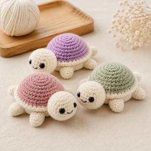 May include: Three small crocheted turtle toys with cream-colored heads and limbs. Each turtle features a distinctively colored shell in soft lavender, dusty pink, and sage green. The turtles have black bead eyes and embroidered smiles.
