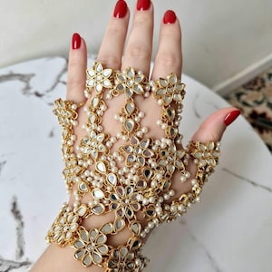 May include: A gold-toned hand harness featuring a series of interconnected flower-shaped motifs inlaid with reflective mirror pieces and accented with small white beads. The jewellery piece extends from a wrist cuff to individual finger rings.