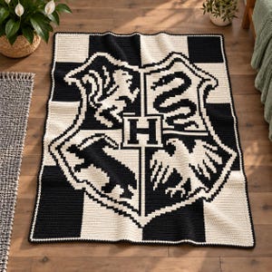 May include: A rectangular crochet blanket featuring a black and white chequered border and a large central crest design. The crest displays a lion, a snake, a badger, and an eagle surrounding a central letter H, all rendered in black and white.