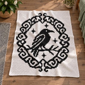 Op de afbeelding: Een rechthoekige witte gehaakte deken met een grote zwarte raaf die in het midden op een tak zit. De vogel is omsloten door een ingewikkelde zwarte decoratieve ovale lijst met krulpatronen en vier kleine vierpuntige sterren als accenten.
