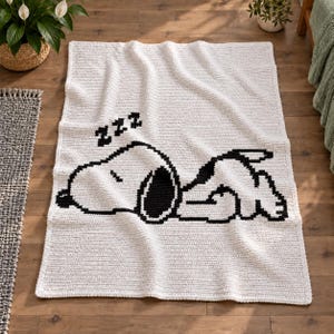May include: A rectangular white crochet blanket featuring a black graphic design of a sleeping dog character lying down with three black Zs above its head. The blanket has a textured, woven appearance and is laid out flat on a wooden floor.