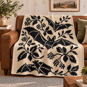 May include: A cream-coloured crochet blanket featuring a repeating pattern of black bats and botanical elements including leaves and small flowers. The textile has a visible stitched texture and a rectangular shape with clean, finished edges.