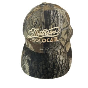 May include: A baseball cap featuring a brown, green, and black camouflage pattern. The front panel displays the embroidered text Mathews in a cursive font above the word SOLOCAM in a block font, both in a light tan thread color.