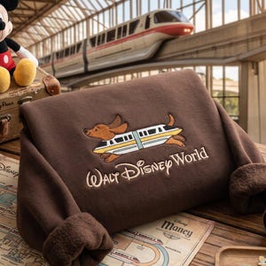 May include: A brown long-sleeve sweatshirt featuring an embroidered graphic of a dachshund riding a monorail train above the text Walt Disney World. The sweatshirt has a soft, plush interior lining and folded cuffs, showcasing a detailed design.