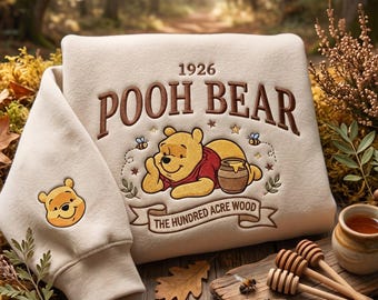 May include: A cream-colored sweatshirt featuring an embroidered graphic of Pooh Bear, a honey pot, and the text 1926 POOH BEAR THE HUNDRED ACRE WOOD. The sleeve displays a small embroidered Pooh Bear face. The design includes small bees and stars.