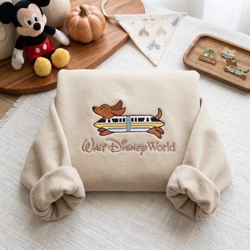 May include: A beige sweatshirt featuring an embroidered graphic of a brown dachshund dog wearing a yellow and white monorail train car around its body. Below the graphic, the text Walt Disney World is embroidered in a brown script font.