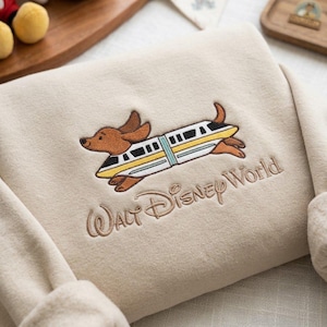 May include: A beige sweatshirt featuring an embroidered design of a brown dachshund dog integrated with a white, yellow, and blue monorail train. Below the graphic, the text Walt Disney World is embroidered in a brown cursive script font.