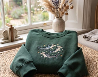 May include: A dark green crewneck sweatshirt featuring an embroidered design of three hammerhead sharks swimming in a circular formation, accented with small yellow stars and white dots, crafted with long sleeves and a ribbed collar and cuffs.