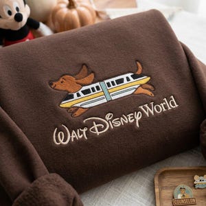 May include: A dark brown sweatshirt featuring an embroidered design of a brown dachshund running alongside a yellow and white monorail train. Below the graphic, the text Walt Disney World is embroidered in a cream-colored script font.