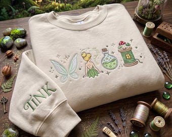 May include: A beige crewneck sweatshirt featuring embroidered designs of fairy wings, a small fairy figure, a potion bottle, and a mushroom-topped spool of thread. The sleeve is embroidered with the green text TINK in a serif font.
