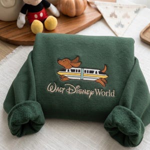 May include: A dark green long-sleeve sweatshirt featuring an embroidered graphic of a brown dachshund dog wearing a white and yellow monorail train as a costume. The text Walt Disney World is embroidered in light tan thread below the graphic.
