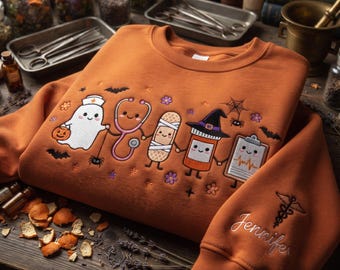 May include: An orange sweatshirt featuring embroidered cartoon medical characters including a ghost nurse, a stethoscope, a bandage, a pill bottle with a witch hat, and a clipboard. The sleeve is embroidered with the name Jennifer and a caduceus symbol.
