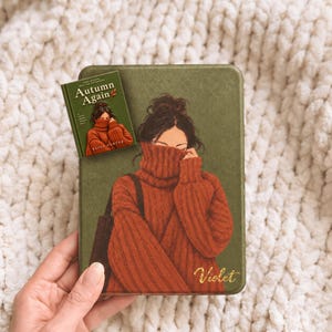 May include: A rectangular olive green e-reader case featuring an illustration of a person in a rust-coloured turtleneck sweater covering their face, with the name Violet in gold script and a small book titled Autumn Again by Elise Carter.