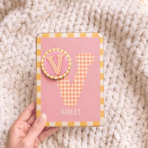 Peut inclure: Une boîte métallique rectangulaire avec un fond rose et une bordure à carreaux jaunes et blancs. Elle présente une grande lettre V à carreaux orange et blancs, une épinglette circulaire avec un V assorti et le nom VIOLET en lettres blanches.