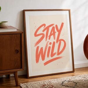 Peut inclure: Une affiche murale encadrée rectangulaire présentant le texte STAY WILD écrit dans un style pinceau audacieux avec de la peinture orange corail sur un fond crème clair. Le cadre est en bois ton moyen avec une bordure passe-partout blanche.