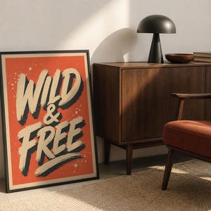 May include: A rectangular framed wall art print featuring a vibrant orange background with the text WILD & FREE written in large, bold, cream-colored brushstroke lettering with dark gray drop shadows and small white paint splatter accents.