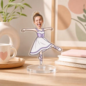 May include: A clear acrylic standee featuring a cartoon-style ballerina illustration with a photographic face cutout. The figure wears a white and lavender dress with lace-up ballet slippers, posed with arms outstretched and one leg extended outward.