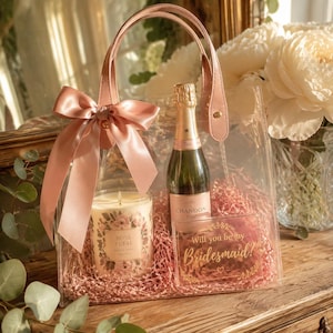 May include: A clear plastic tote bag with pink handles and a large pink satin bow. Inside is a bottle of Chandon sparkling wine, a Rose Petal scented candle in a glass jar, and a rectangular acrylic sign with the text Will you be my Bridesmaid?