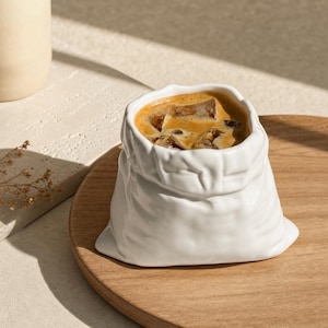 May include: A white ceramic mug shaped like a crumpled fabric sack filled with iced coffee. The exterior features a textured, folded appearance mimicking a soft bag, while the interior holds a dark brown liquid with several visible ice cubes.