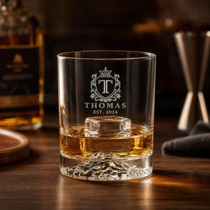 May include: A clear glass tumbler containing amber liquid and a single ice cube. The glass features an etched crest with the letter T, the name THOMAS, and the text EST. 2024. The base of the glass has a raised, textured mountain peak design.