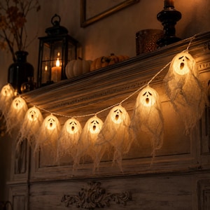 Può includere: Una catena di luci LED con coperture a forma di fantasma in tessuto bianco e tratti del viso neri. Ogni fantasma è illuminato dall'interno, emettendo una luce calda. La catena è drappeggiata orizzontalmente su una mensola in legno.