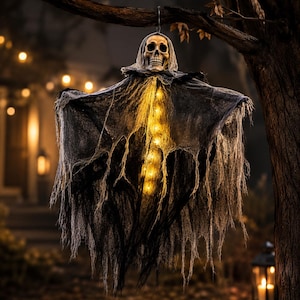 Può includere: Una decorazione di Halloween sospesa con una testa di teschio umano realistica drappeggiata in un tessuto grigio scuro logoro. La figura è illuminata dall'interno da una serie verticale di luci LED color giallo caldo.