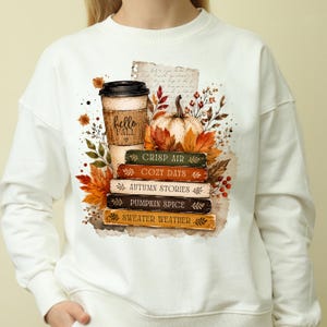 Puede incluir: Una sudadera de cuello redondo color crema con un estampado gráfico de una taza de café con la etiqueta "hello FALL" sobre una pila de cinco libros titulados "CRISP AIR", "COZY DAYS", "AUTUMN STORIES", "PUMPKIN SPICE" y "SWEATER WEATHER" con calabazas y hojas.