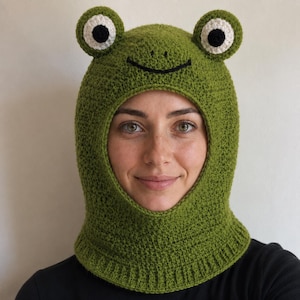 May include: A green crochet balaclava featuring a frog design with two protruding eyes on top, each with a white and black center. The hat has a stitched black mouth and a ribbed bottom edge, covering the head and neck with a circular face opening.
