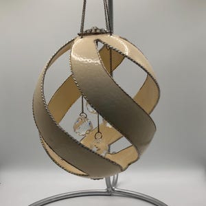 May include: A spherical ornament featuring cream-colored curved bands with silver-toned braided edges. The structure is suspended by a thin chain and contains multiple clear, faceted crystal-like beads hanging from the center of the open frame.