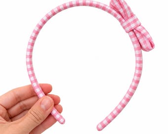 May include: A pink and white gingham patterned headband featuring a fabric-covered band and a matching fabric bow attached to the right side. The headband has a rounded shape and is constructed from a checkered textile material.
