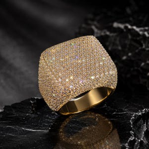 Peut inclure: Une bague dorée dotée d'une large surface supérieure carrée entièrement recouverte de nombreuses petites pierres claires et scintillantes. L'anneau de la bague est lisse et poli, présentant une finition métallique dorée réfléchissante.