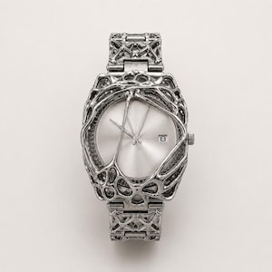 Peut inclure: Une montre-bracelet argentée dotée d'un cadran circulaire avec un affichage de la date à trois heures. Le boîtier et le bracelet de la montre sont entourés d'une structure métallique complexe, organique et réticulée avec une finition polie.