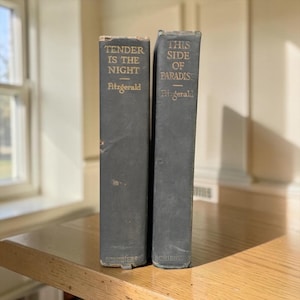 May include: Two vintage hardcover books with dark grey spines standing upright. The left book is titled Tender Is The Night by Fitzgerald, and the right book is titled This Side Of Paradise by Fitzgerald. Both spines feature gold lettering and the publisher name Scribners at the bottom.
