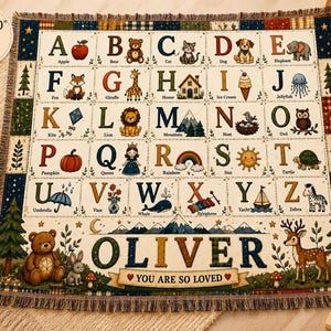 Könnte beinhalten: Eine gewebte Decke mit den Maßen 203 cm mal 152 cm, die ein Alphabet-Gitter mit Illustrationen für jeden Buchstaben von A bis Z zeigt. Der untere Bereich zeigt den Namen OLIVER in großen Buchstaben mit dem Text YOU ARE SO LOVED auf einem Banner.