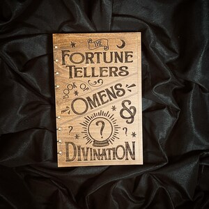 May include: A rectangular wooden book cover with a dark brown finish, featuring laser-engraved text that reads The Fortune Tellers Book of Omens & Divination. The design includes a central crystal ball icon, stars, a crescent moon, and small metal rings.