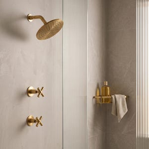 May include: A brushed gold shower system featuring a round shower head, two cross-handle valves, and a corner shelf holding a clear bottle with amber liquid and a beige textured washcloth, all mounted against light grey stone-look wall tiles.