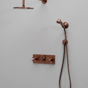 May include: A wall-mounted shower system in a copper finish, featuring a circular overhead rain shower head, a handheld shower wand with a flexible hose, and a rectangular control plate with two cross-handle valves and a central diverter.